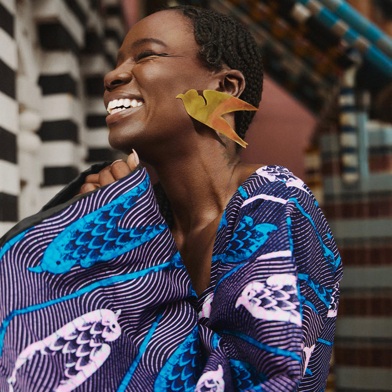Sarah Diouf wearing vibrant Vlisco fabric with bird motifs, smiling joyfully and styled with bold statement earrings in an African fashion photoshoot.