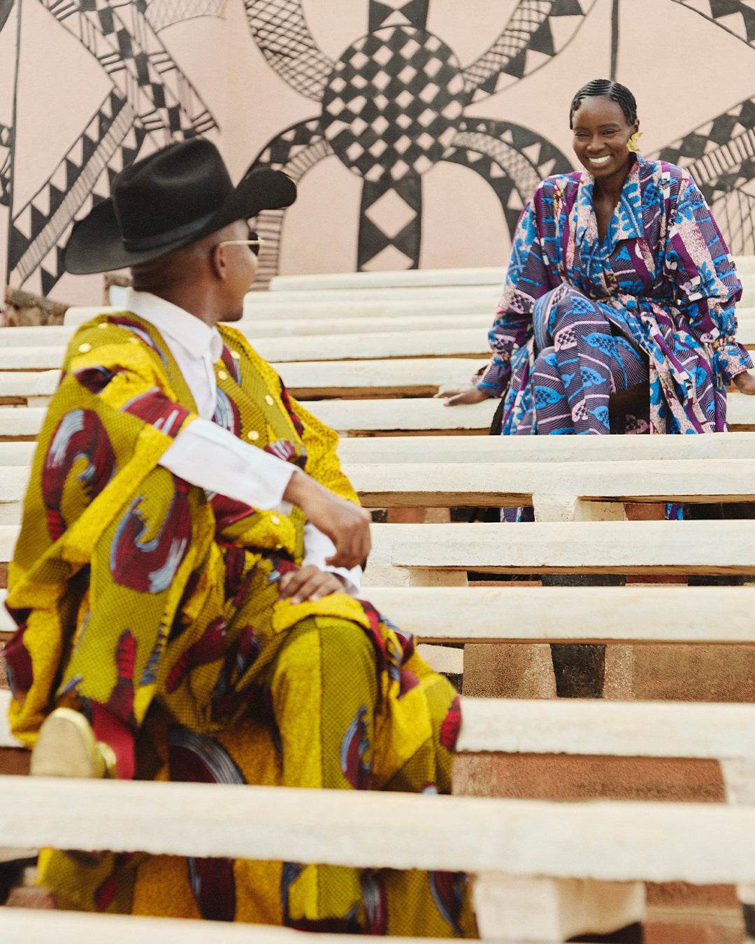 Trevor Stuurman and Sarah Diouf wearing vibrant Vlisco designs, seated on outdoor steps with a geometric mural backdrop, celebrating African fashion and heritage.