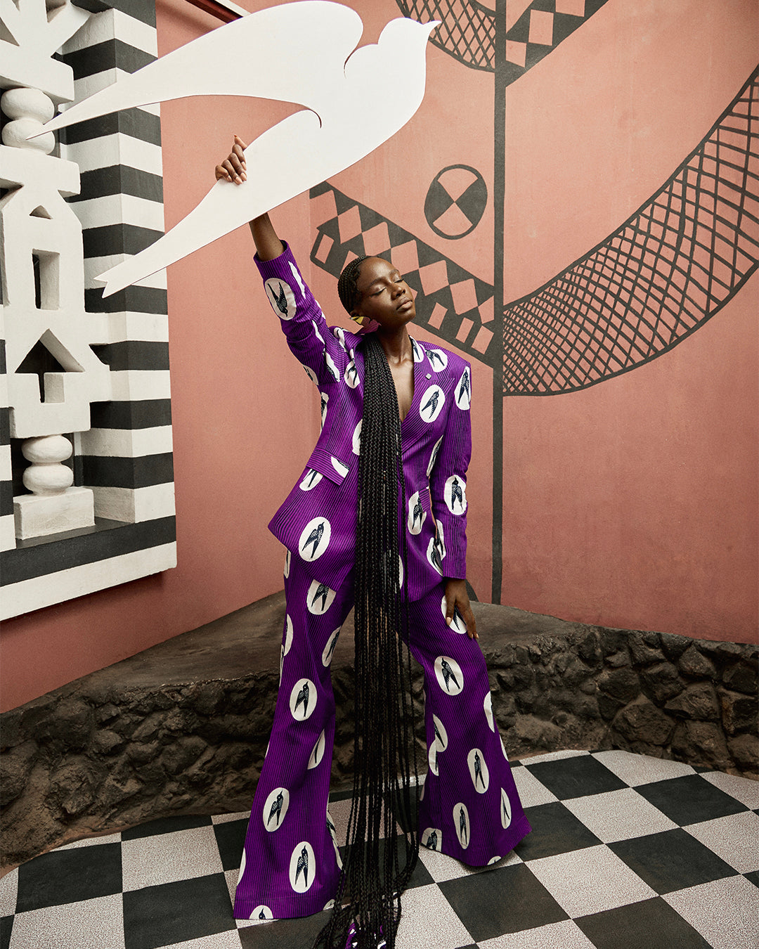 Sarah Diouf in a tailored purple Vlisco suit of the speed bird design, holding a large dove prop against a graphic wall in an artistic fashion portrait