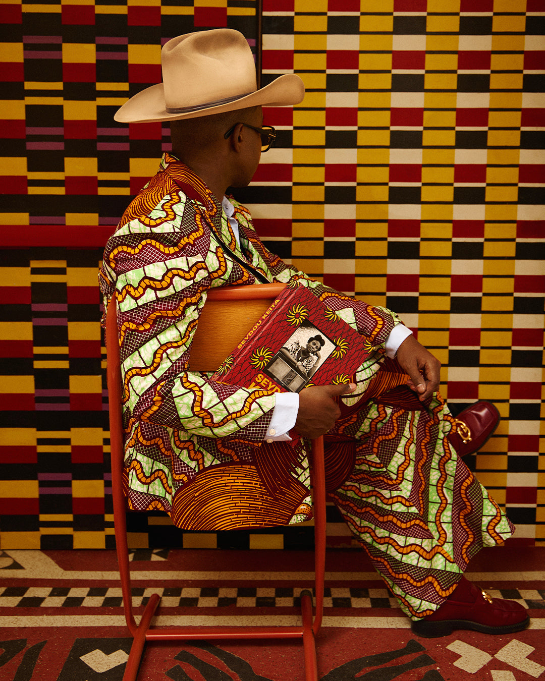 Trevor Stuurman dressed in a colorful Vlisco suit and cowboy hat, seated against a geometric wall while holding a Seydou Keïta book, blending fashion and culture.