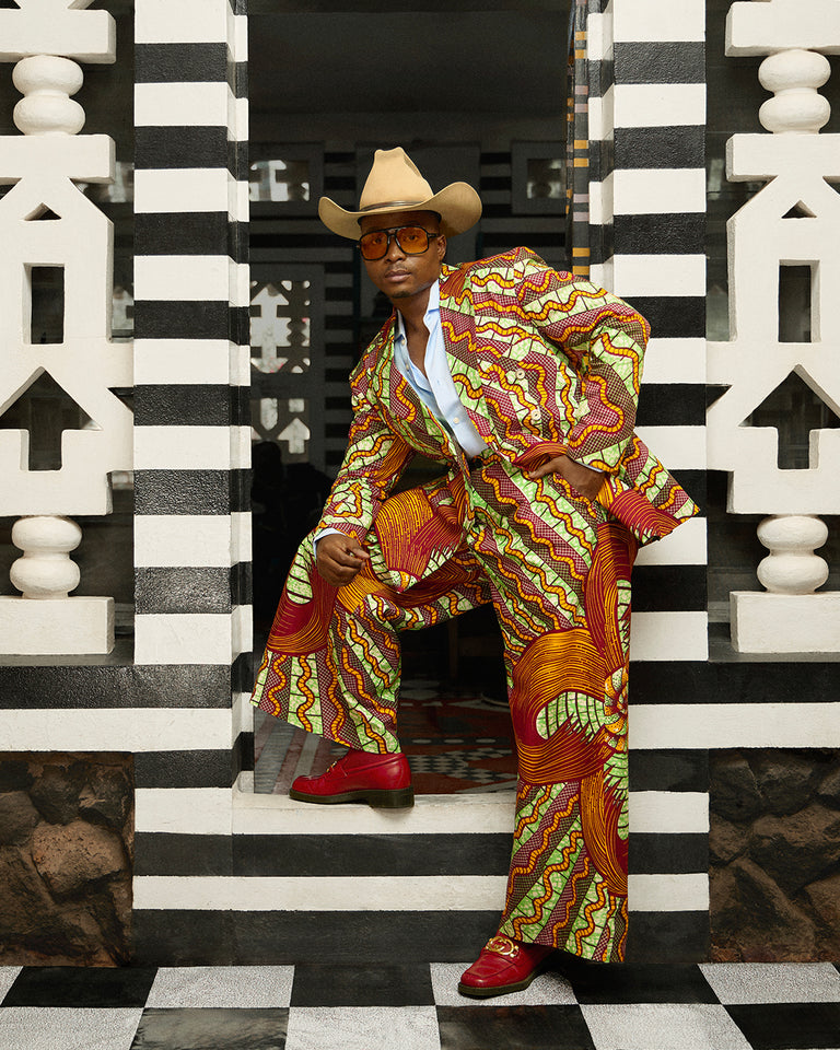 Trevor Stuurman wearing a bold Vlisco suit in red, green, and yellow patterns, styled with a cowboy hat and sunglasses, posing against a black-and-white striped architectural backdrop